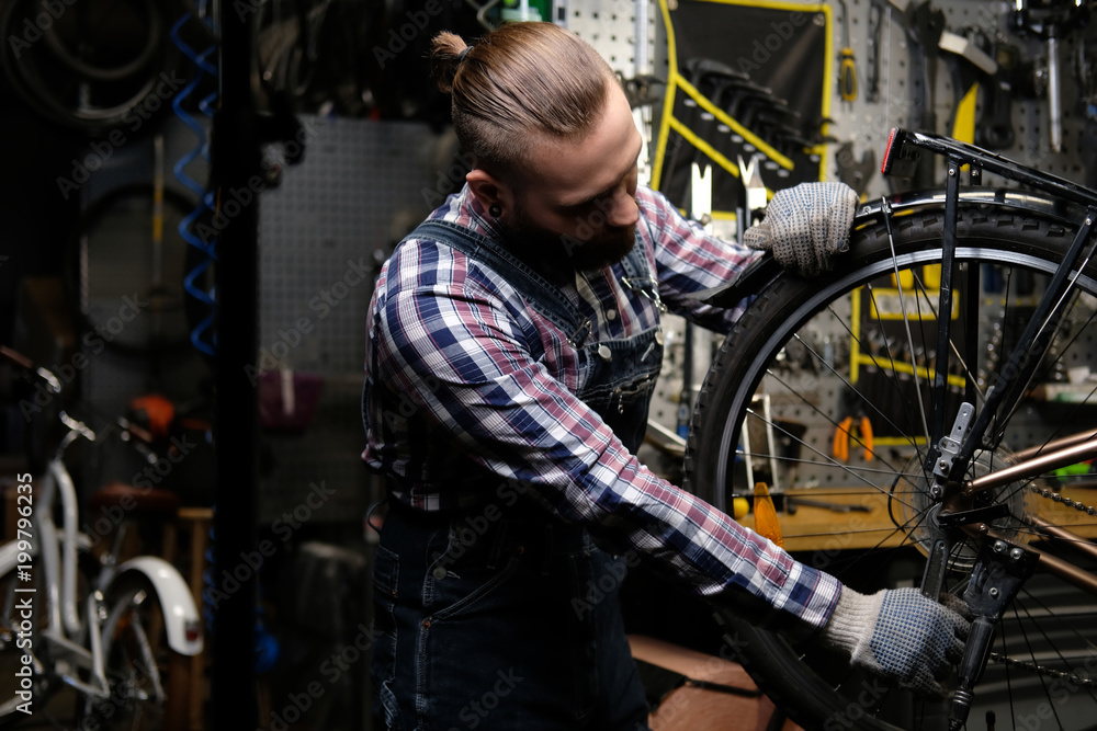 Handsome stylish male wearing a flannel shirt and jeans coverall, working with a bicycle wheel in a repair shop. A worker using a wrench mounts the wheel on a bike in a workshop.