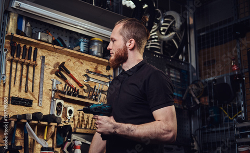 Handsome bearded redhead worker with haircut, holding an electric screwdriver, working in a repair shop. 