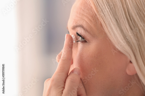 Senior woman putting contact lens in her eye on blurred background