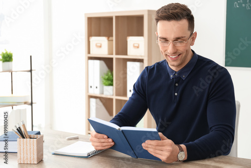 Young male teacher with book sitting at table in classroom