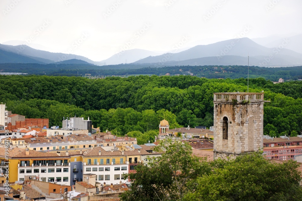 View of the city of Girona from the medieval pedestrian border wall. Roofs of houses, trees, mountains in a haze in the background. Cloudy sky. Girona, Spain View of the city of Girona from the medieval pedestrian border wall. Roofs of houses, trees, mountains in a haze in the background. Cloudy sky. Girona, Spain