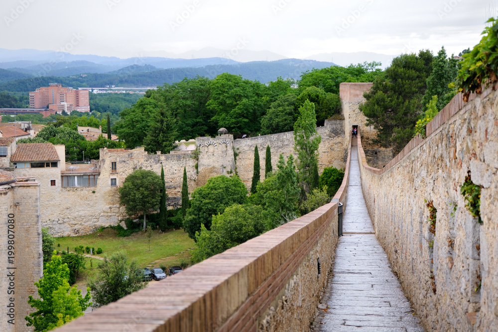 Fototapeta premium View of the city of Girona from the medieval pedestrian border wall. Roofs of houses, trees, mountains in a haze in the background. Cloudy sky. Girona, Spain