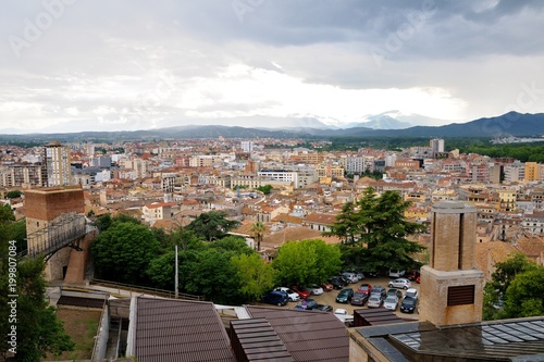 Fototapeta Naklejka Na Ścianę i Meble -  View of the city of Girona from the medieval pedestrian border wall. Roofs of houses, trees. Storm clouds over the city, somewhere sunlight makes its way through the clouds. Girona, Spain