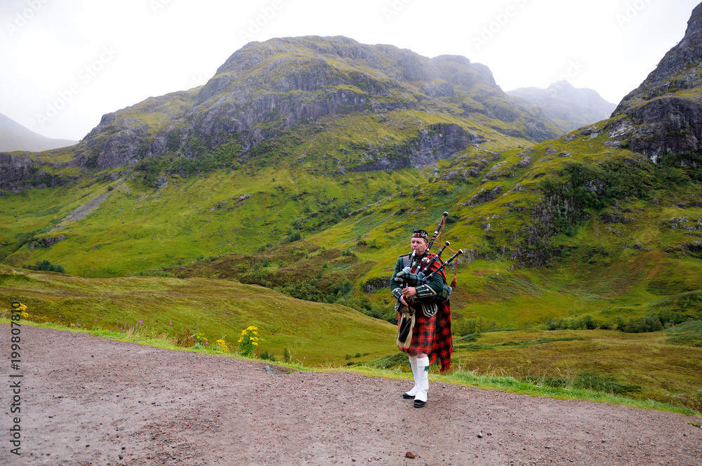 Piper in traditional Scottish outfit plays on bagpipes in Scottish Highlands. In the background of the mountain. Cloudy autumn day.