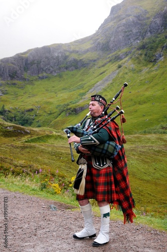 Slika na platnu Piper in traditional Scottish outfit plays on bagpipes in Scottish Highlands