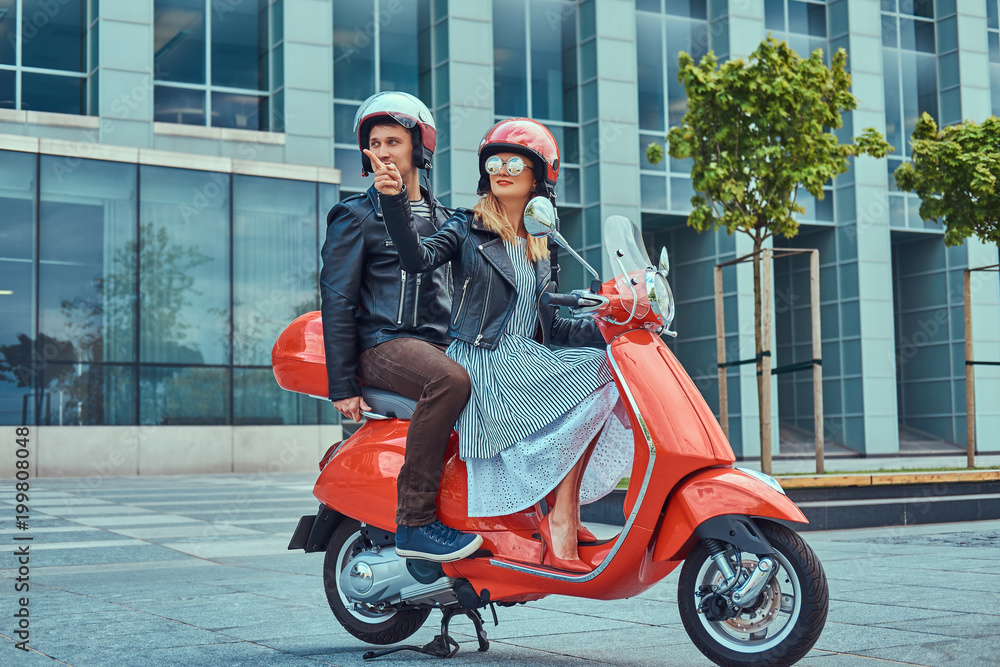 Attractive happy couple, a handsome man and sexy female riding together on a red retro scooter in a city.