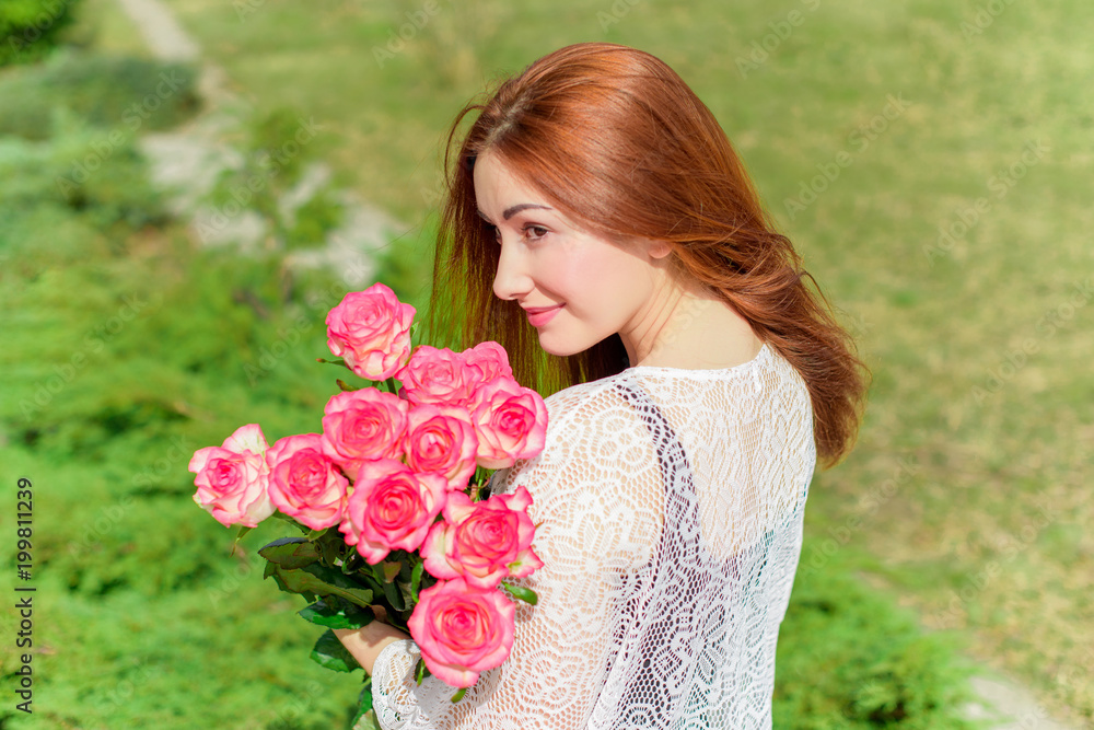 Pretty girl in a tender knitted dress, holding her hands on her shoulder a bouquet with pink roses on the background of garden 