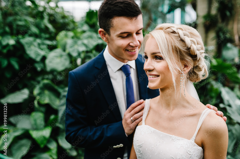 Portrait the groom in wedding suit and the bride in dress are standing and hugging in the Botanical green garden full of greenery. Wedding ceremony. Portrait the groom in wedding suit and the bride in dress are standing and hugging in the Botanical green garden full of greenery. Wedding ceremony.