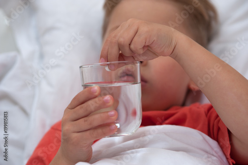 Little sick cute cboy holding a pill and a glass of water lying on the bed