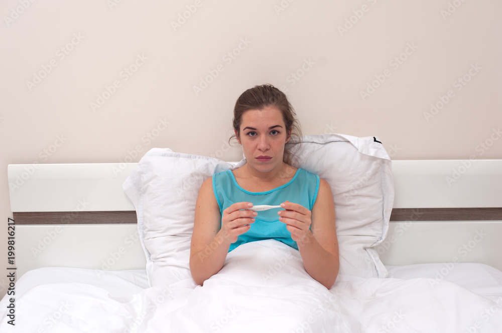 Disease and poor health. Young woman in blue dress is sitting on the bed and holding thermometer in hands looking at the camera Disease and poor health. Young woman in blue dress is sitting on the bed and holding thermometer in hands looking at the camera