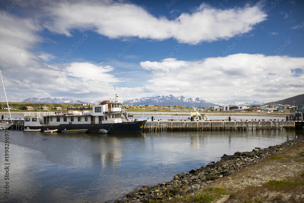 The clouds and mountains reflect off the waters of Ushuaia, Argentina while an old boat sits docked for life in the foreground.