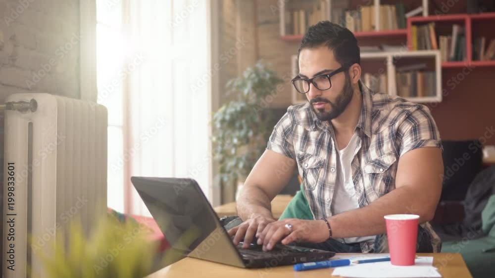 Handsome bearded man working at the computer, busy boy in stylish glasses typing quickly on black device, wearing fashionable checked shirt, sitting on comfortable green chair Handsome bearded man working at the computer, busy boy in stylish glasses typing quickly on black device, wearing fashionable checked shirt, sitting on comfortable green chair