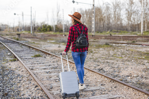 young beautiful woman wearing casual clothes, walking by the railway with suitcase and a bag. She is smiling. Outdoors lifestyle. Travel concept.