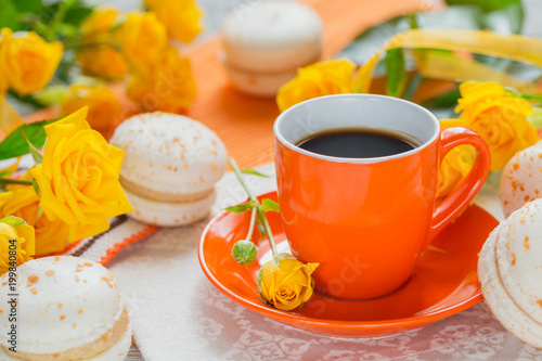 Orange cup of black coffee, yellow roses flowers and sweet pastel french macaroons