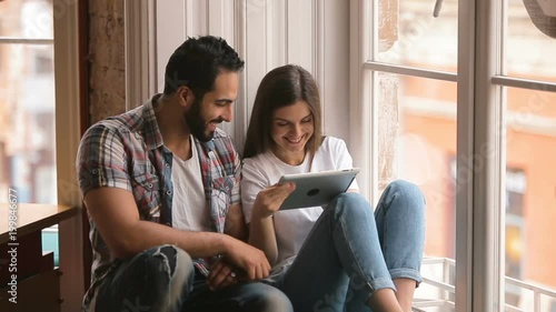 Joyful couple having fun with tablet, well-built man sliding on the gadget screen, smiling brunette holding tablet nearby, playing computer games near big cafe windows during coffee time