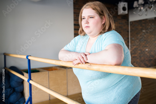 Waist up portrait of obese young woman standing at bar in dance class looking pensively at window, copy space photo