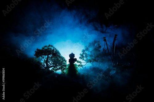 Horror Halloween decorated conceptual image. Alone girl with the light in the forest at night. Silhouette of girl standing between trees with surreal light . Selective focus.