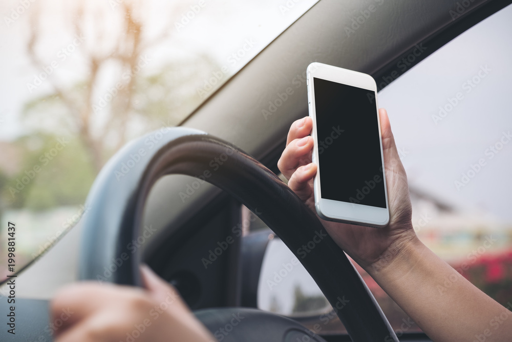 Mockup image of a hand holding and using white mobile phone with blank black desktop screen while driving car on the road