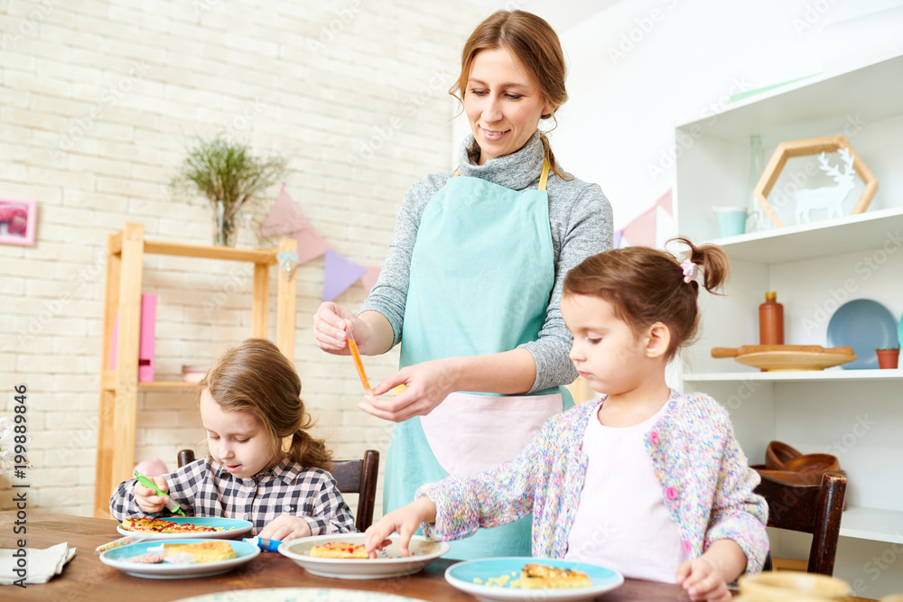 Attractive young woman wearing apron teaching her little daughters to decorate waffles with colorful glaze while spending day at home