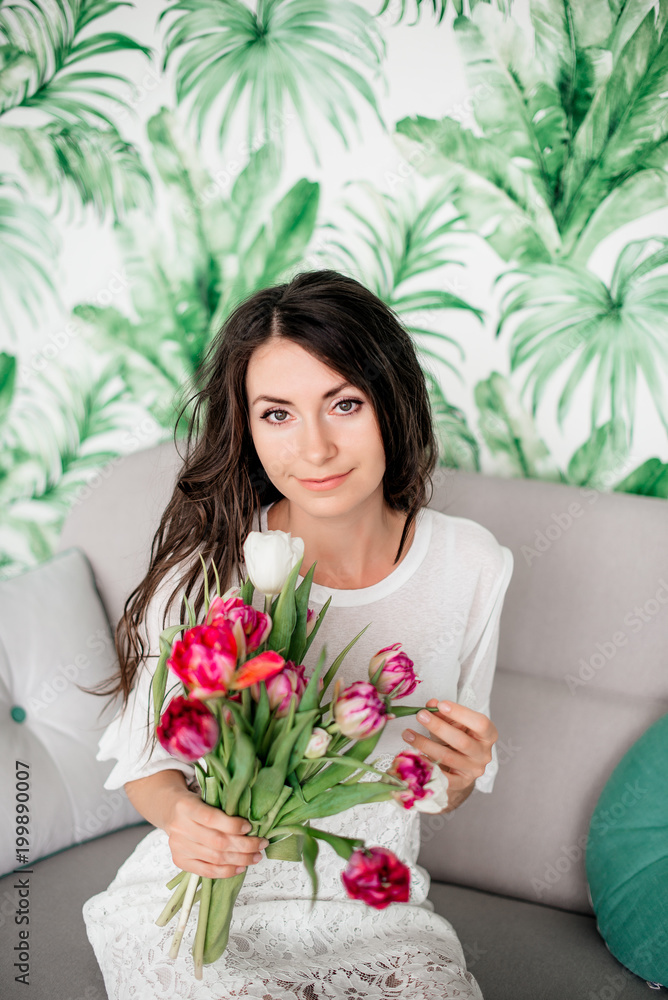 Beautiful girl with dark hair sitting on a sofa in a white dress and holding a bouquet of tulips. Green background