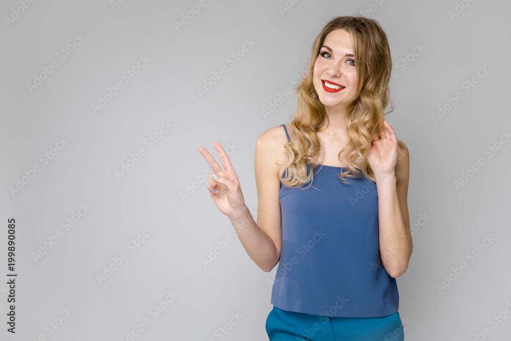 Charming young blonde girl in blue clothes smiling with victory gesture on gray background Charming young blonde girl in blue clothes smiling with victory gesture on gray background