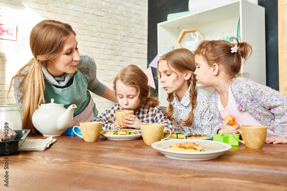 Cute fair-haired girl drinking hot tea from cup while her sisters and mother looking at her with curiosity, appetizing pastry waiting for them, interior of dining room on background Cute fair-haired girl drinking hot tea from cup while her sisters and mother looking at her with curiosity, appetizing pastry waiting for them, interior of dining room on background