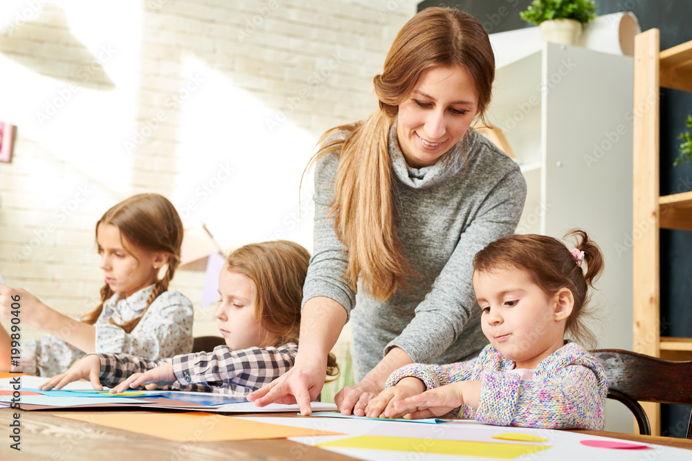 Working process at art studio: little pupils sitting at wooden desk and wrapped up in greeting cards creation from colored paper while their friendly young teacher assisting them