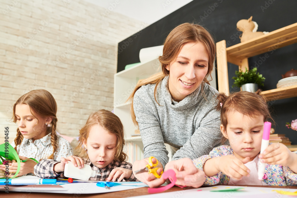 Obraz premium Concentrated little girls sitting at wooden desk of modern art studio and expressing creativity with help of colored paper while their friendly young teacher keeping eye on them.