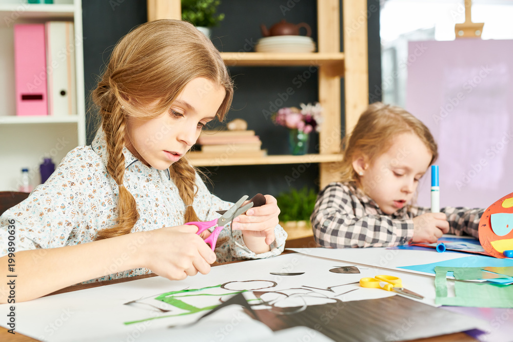 Pretty girl with two braids cutting shape from colored paper while her little sister wrapped up in drawing with felt-tip pens, interior of cozy living room on background Pretty girl with two braids cutting shape from colored paper while her little sister wrapped up in drawing with felt-tip pens, interior of cozy living room on background