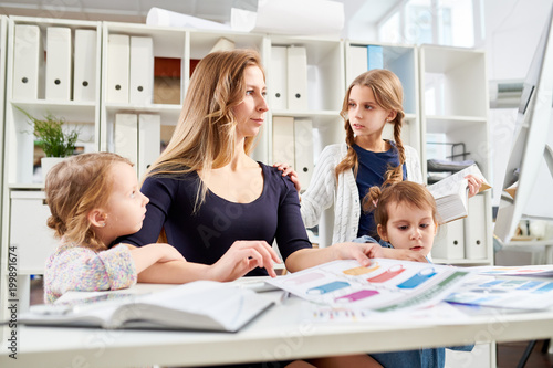 Beautiful young entrepreneur sitting at desk and working on ambitious project while her little children trying to attract her attention, interior of modern office on background
