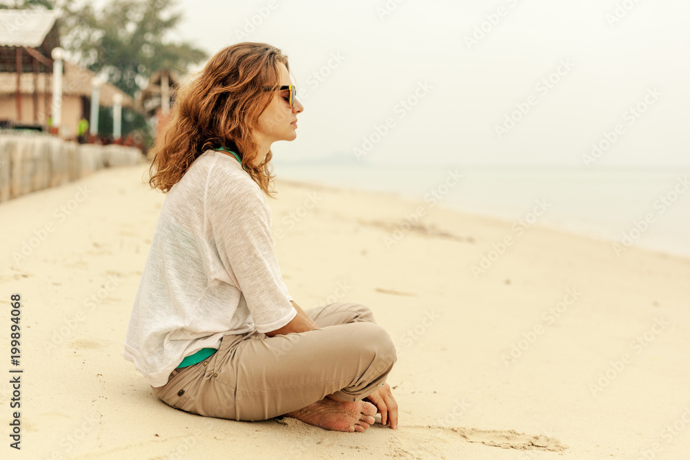 Beautiful young woman with wavy hair sitting on the beach on white sand and admiring the view