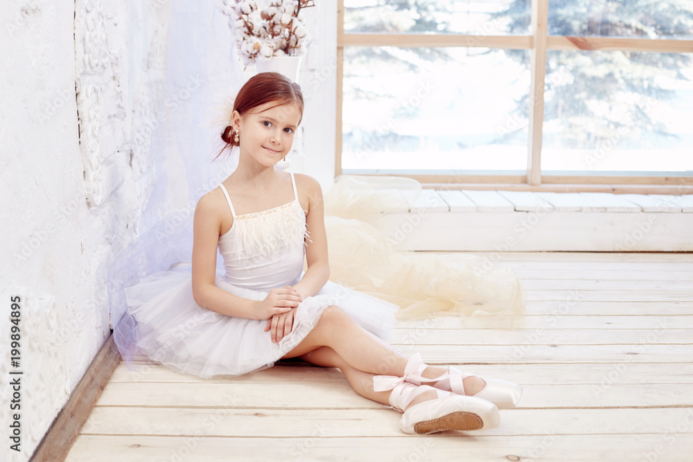 Little prima ballet. Young ballerina girl is preparing for a ballet performance. Girl in a white ball gown and Pointe near the window, beautiful red hair. Young theater actress