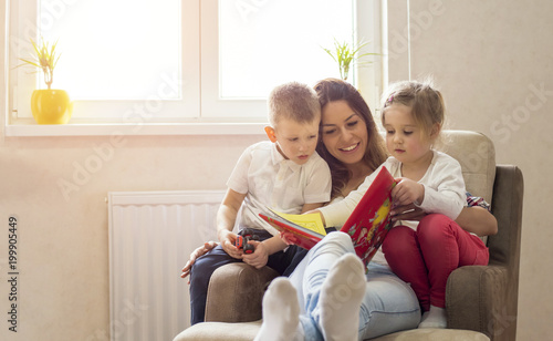 Mother and children reading book and having fun while spending time together at home