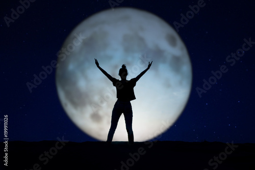 Silhouette of a woman standing in the night with the arms up, giant moon in the background