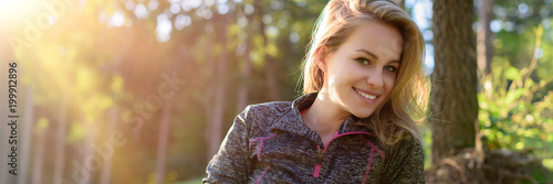 Attractive smiling young blond woman, wearing sportswear, relaxing after workout in a forest. Web banner.