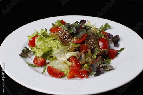 On a white plate salad of tomatoes, fried meat, onions, lettuce, cabbage strewed with sesame seeds, basil, on a black background.Dietary food