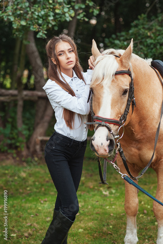 portrait beautiful young girl in white shirt and black pants with beauty long hair next horse in forest. Fashionable elegance woman posing near animal. Beauty Lifestyle Fashion People Animals concepts
