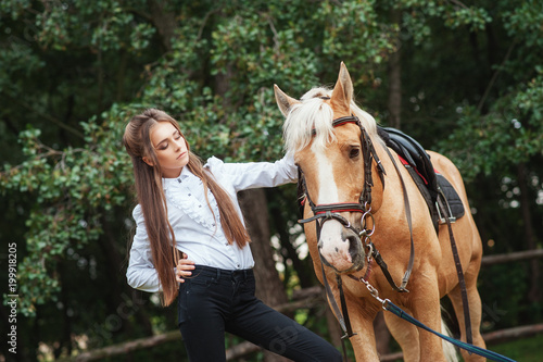 portrait beautiful young girl in white shirt and black pants with beauty long hair next horse in forest. Fashionable elegance woman posing near animal. Beauty Lifestyle Fashion People Animals concepts © victoriazarubina