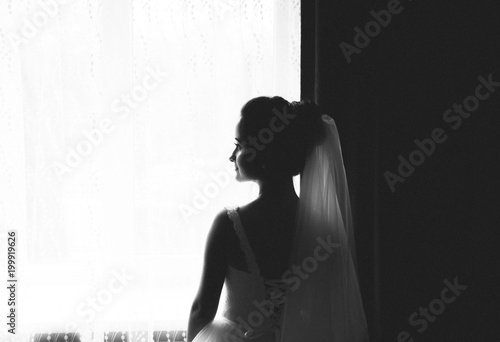 Young gorgeous bride is standing near the window waiting for her groom. Tender light and tulle veil. Beautiful photo of bridal morning. Black and white.
