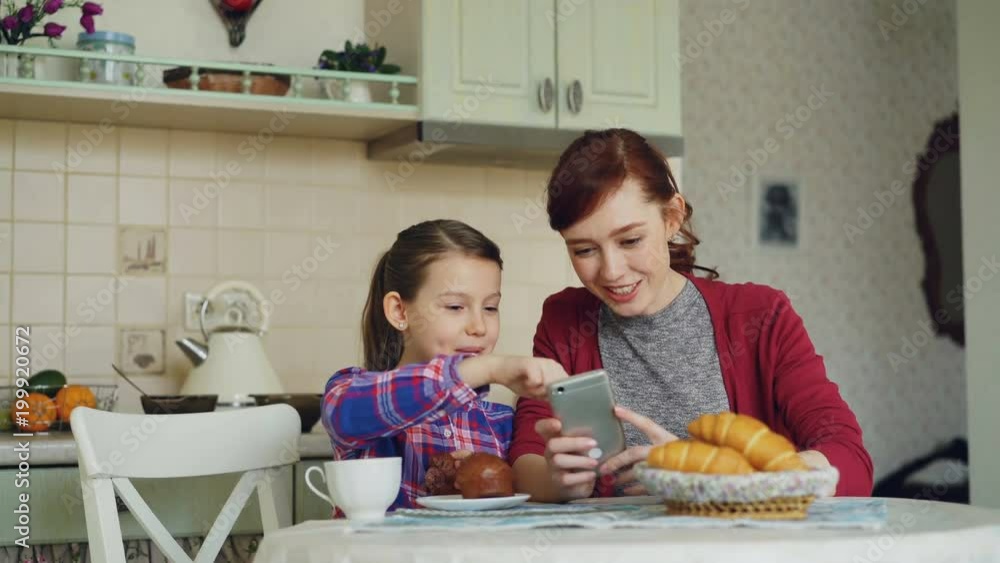Cheerful mother and cute daughter browsing smartphone together and smiling during breakfast in the morning in kitchen at home Cheerful mother and cute daughter browsing smartphone together and smiling during breakfast in the morning in kitchen at home