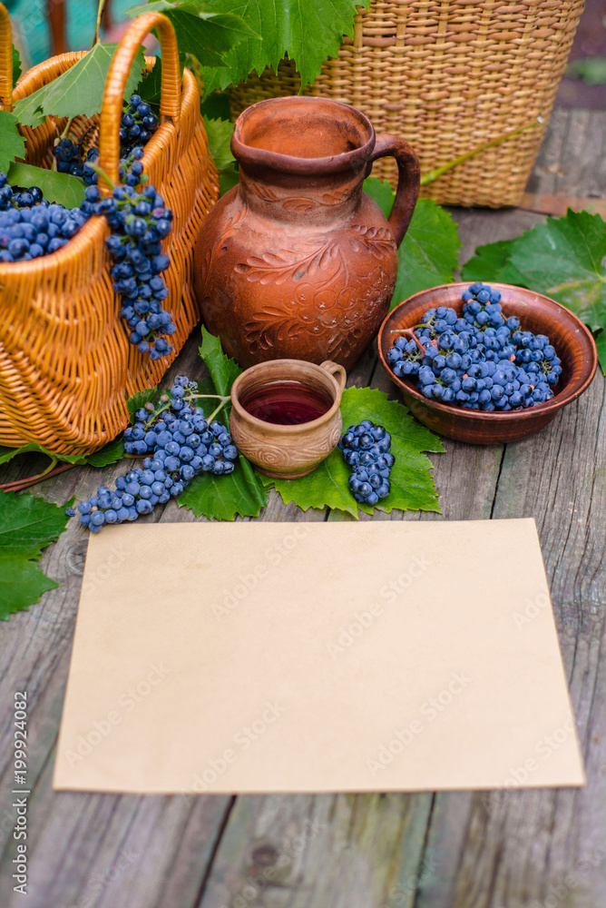 Horizontal sheet of paper lay beside basket,bowl with grapes, jar and cup with wine on rustic wood. Wine making vertical background