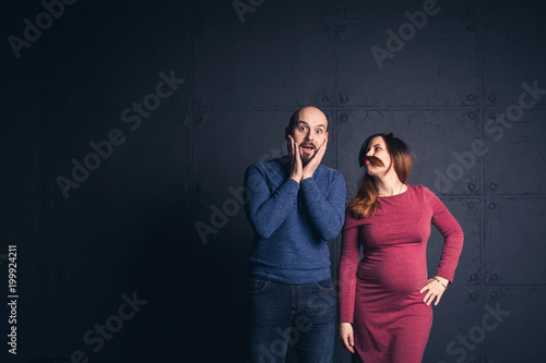 bearded man in shock when pregnant woman makes mustache out of hair in studio on dark background. Copy space.