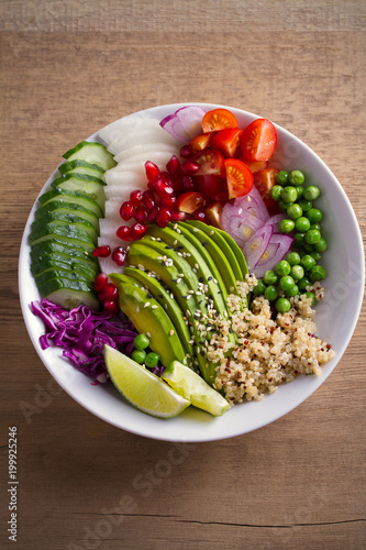 Vegetarian healthy food: quinoa, avocado, pomegranate, tomatoes, green peas, radish, red cabbage and lime salad in bowl on wooden table. overhead, vertical