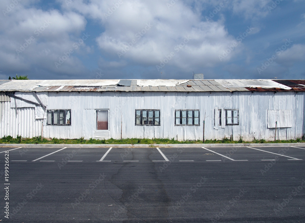 a row of dilapidated abandoned white tin sheet corrugated buildings with empty car park and blue sky and clouds a row of dilapidated abandoned white tin sheet corrugated buildings with empty car park and blue sky and clouds