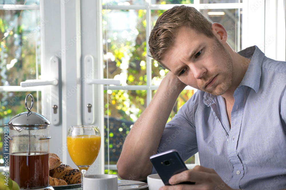 Handsome man sitting at breakfast table with cereal, fruit, flowers, coffee and croissants using his mobile device phone and frowning, staring Handsome man sitting at breakfast table with cereal, fruit, flowers, coffee and croissants using his mobile device phone and frowning, staring