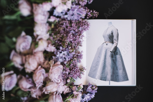 VORONEZH/RUSSIA-02.01.2018: Fashion book depicting old Dior dress with fresh flowers (roses, lilac and mattiolas in lavender color) on the black background, top view, flat lay photo