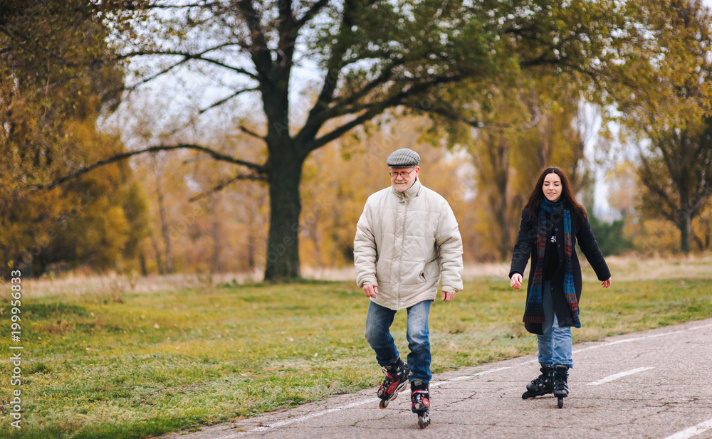 Fototapeta premium Happy old man goes on rollers with his daughter on the road in autumn park. Happy pension. Active old people.