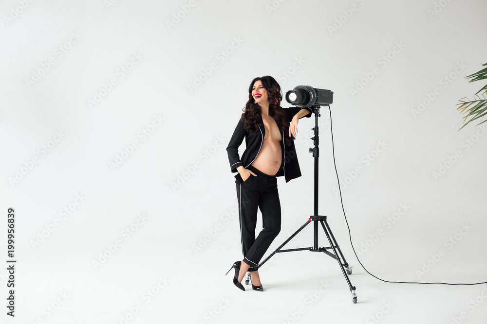 Cheerful photo shoot. Beautiful young pregnant woman in black silk pajama looking away with smile while leaning to lightning tool against white background Cheerful photo shoot. Beautiful young pregnant woman in black silk pajama looking away with smile while leaning to lightning tool against white background