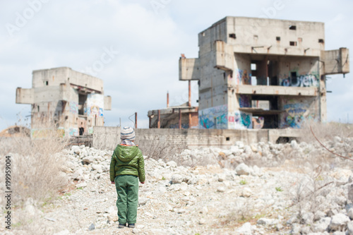 Fototapeta little child in jacket stands against ruins of building as result of war conflic