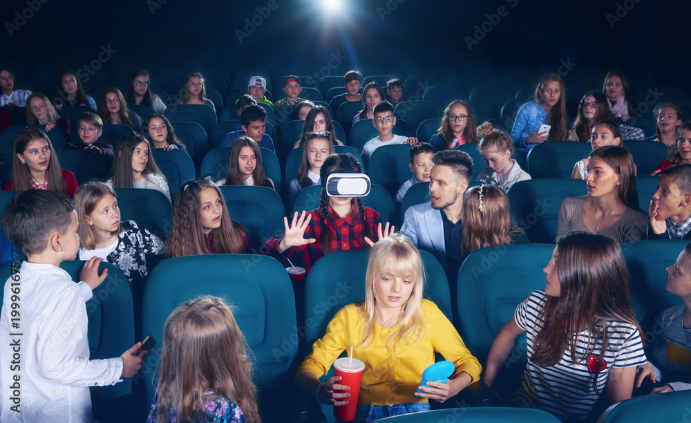 Cinema hall full of young people and children,looking surprised and exited, watching on girl wearing 3D-eyeglasses. People sitting on comfortable plases feeling interested. Cinema hall full of young people and children,looking surprised and exited, watching on girl wearing 3D-eyeglasses. People sitting on comfortable plases feeling interested.